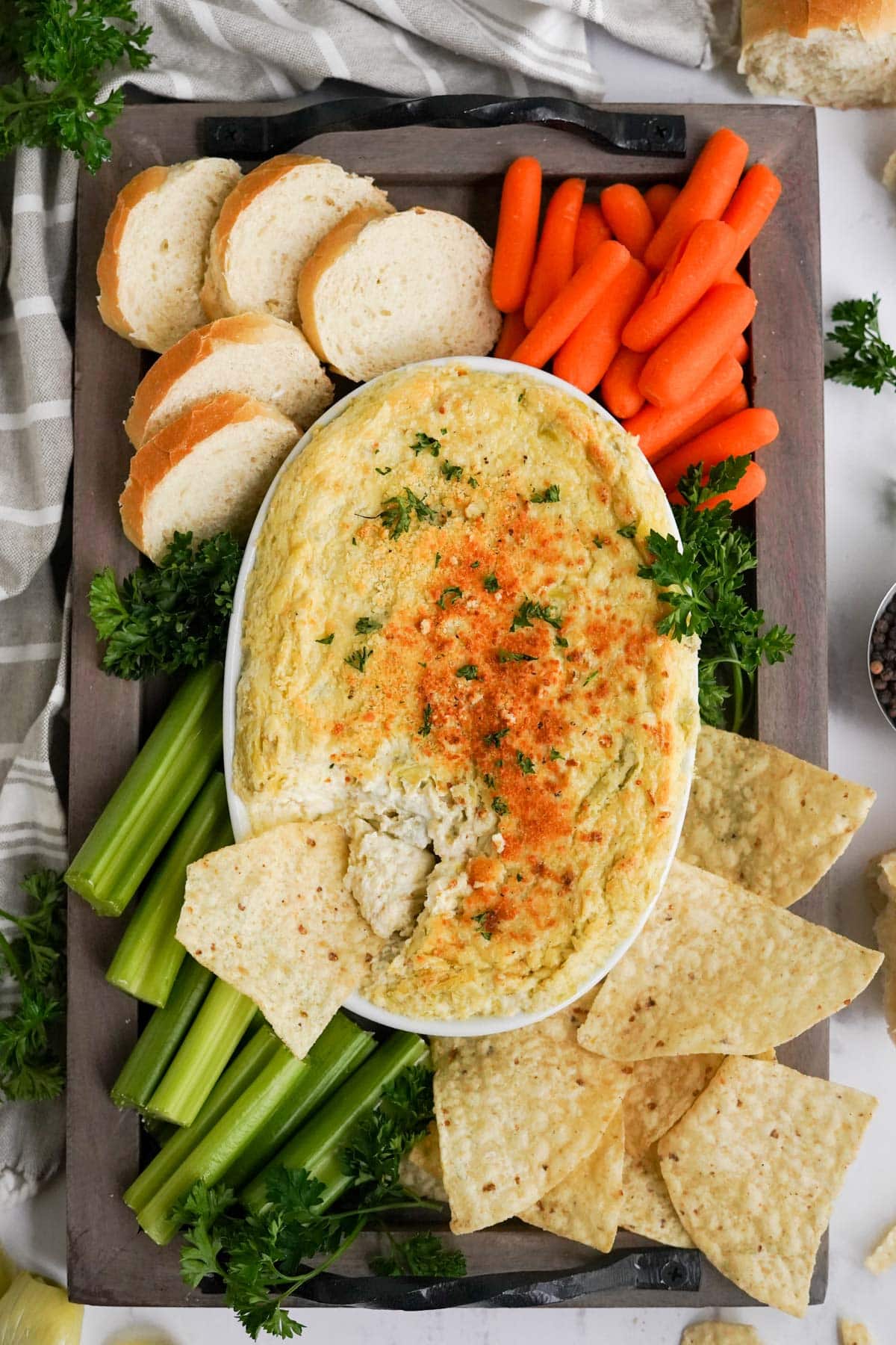 artichoke dip in a white dish with carrots, celery, bread and tortillas chips