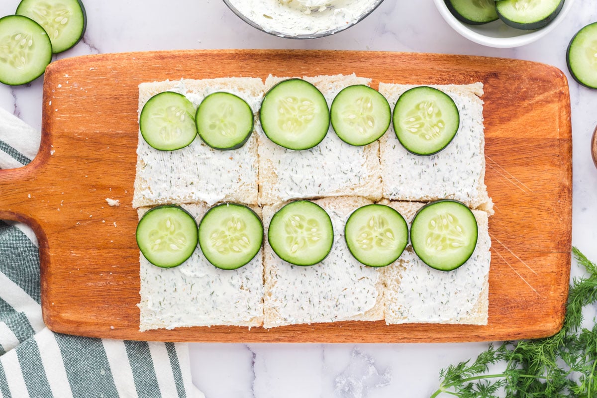 bread, herbed cream cheese and cucumbers on a wood cutting board