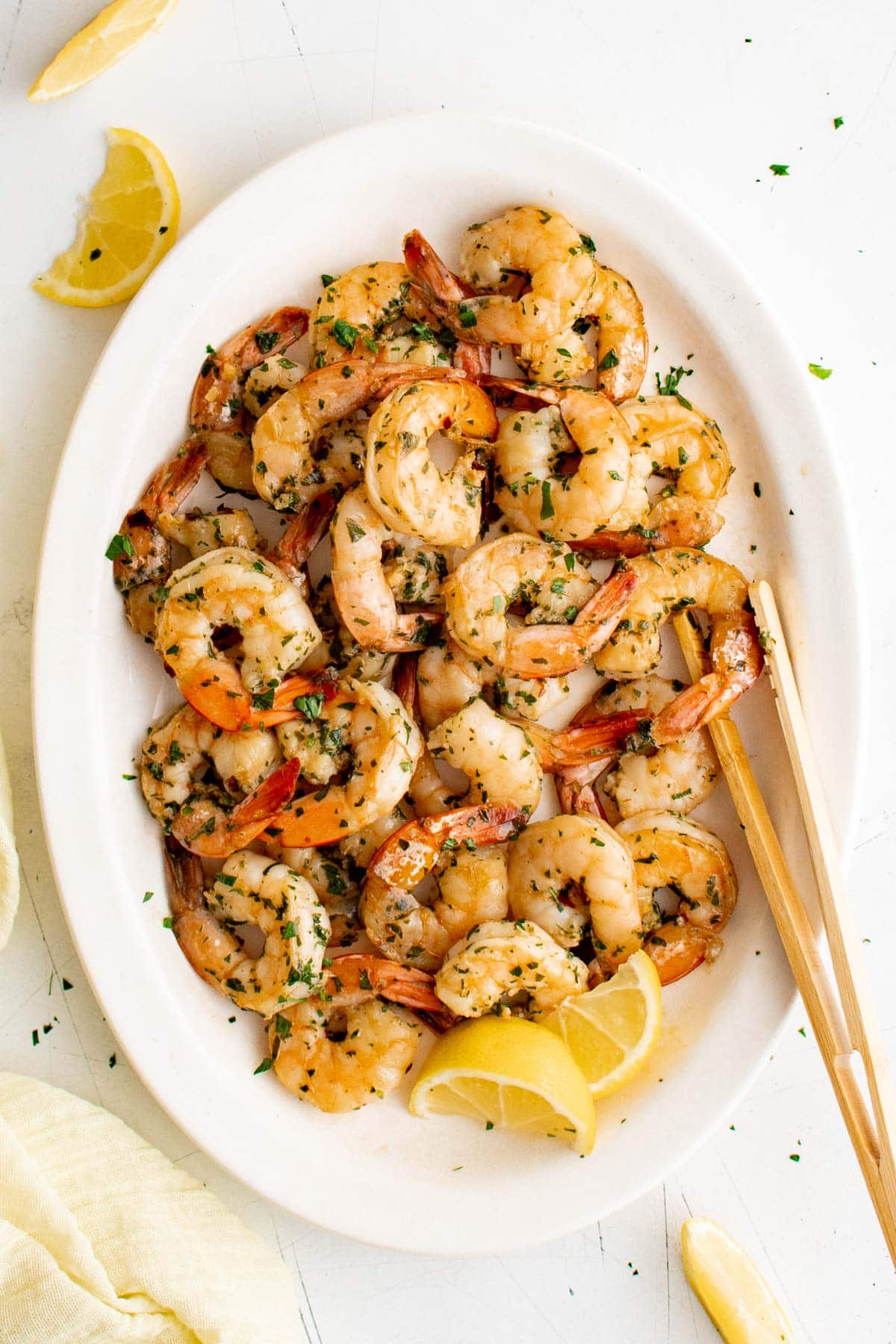 Marinated shrimp on a white platter with lemon slices and wood tongs.
