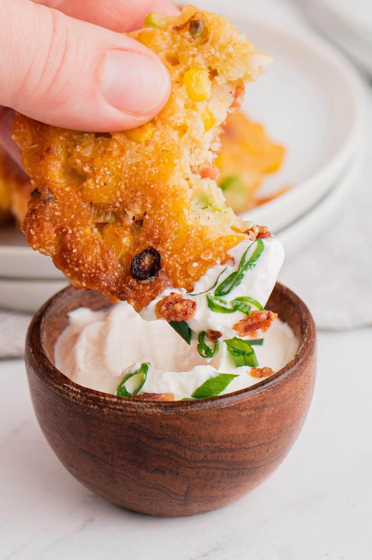 A hand dipping a corn fritter into a sour cream dipping sauce.