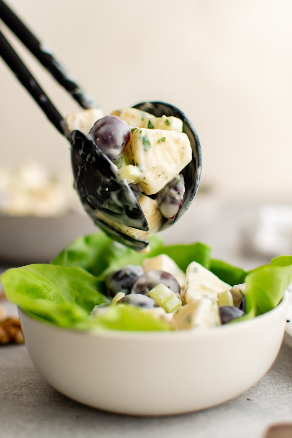 Serving spoons holding a spoonful of waldorf salad and a white bowl with lettuce and salad. 