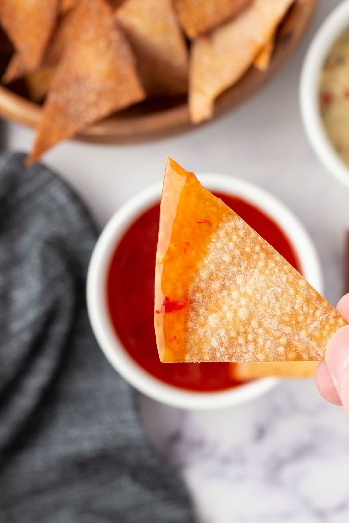 A hand holding a wonton chip over red sauce.