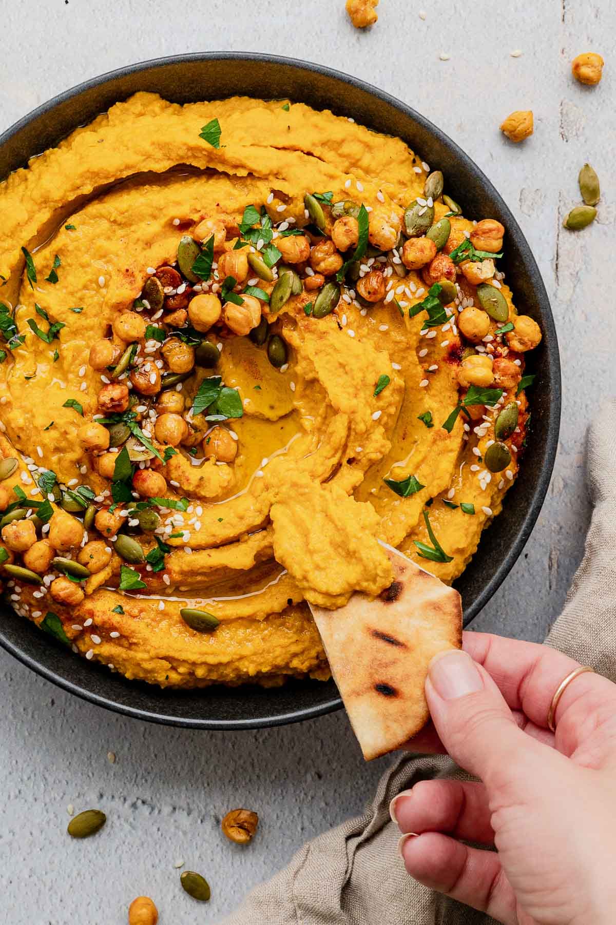 Pumpin hummus topped with chickpeas, parsley and sesame seeds, a hand holds a pita triangle.