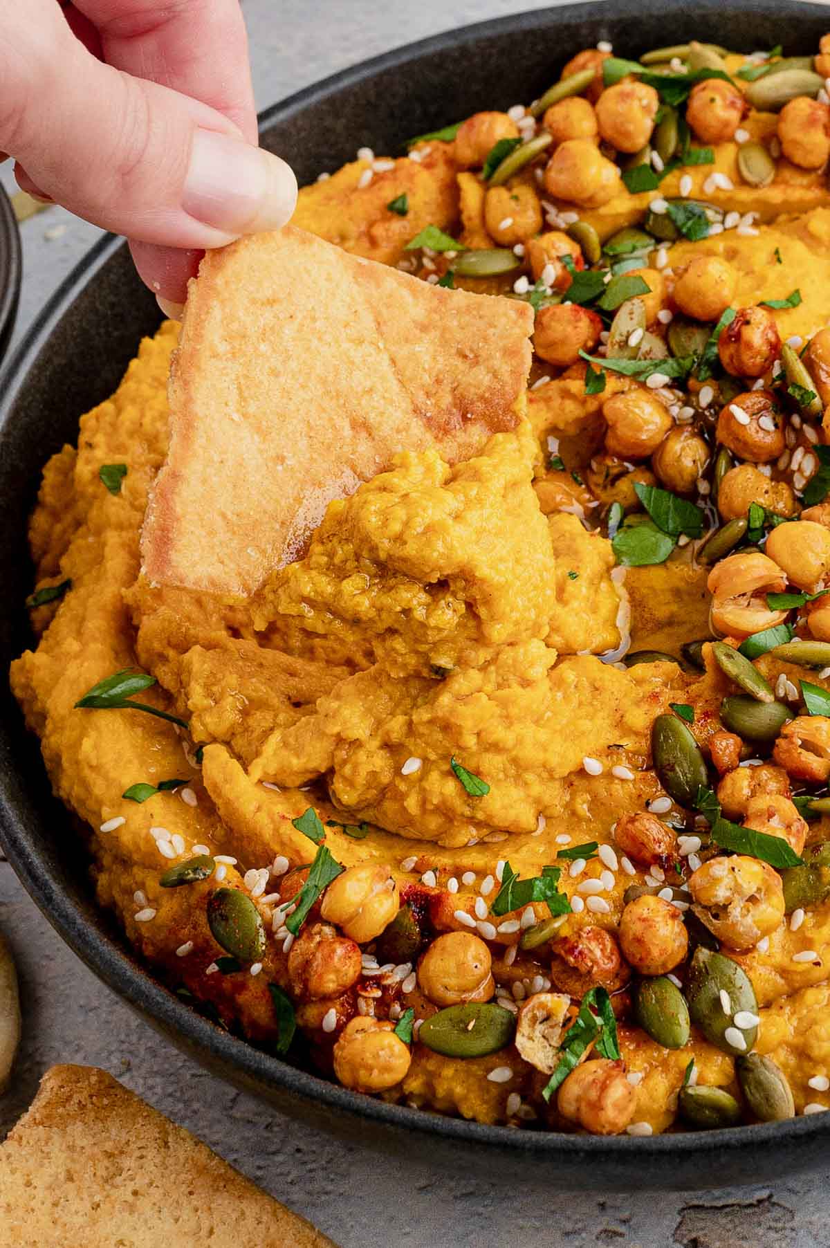 Pumpkin hummus with chickpeas, parsley and a pita chip with a hand.