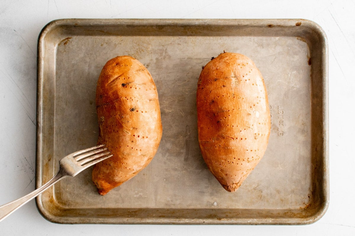 Two sweet potatoes on a baking sheet being pierced with a fork.